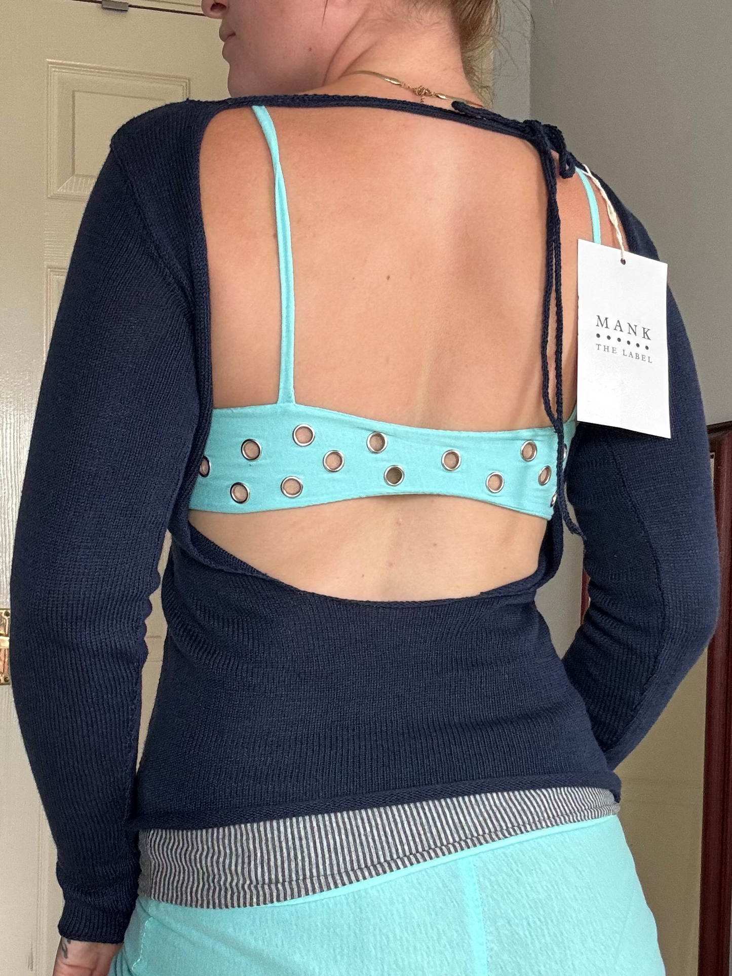Person wearing a blue long-sleeve top with a turquoise bralette underneath, holding a 'MANK' tag.